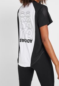 Černé a bílé sportovní tričko s síťovými rukávy, s velkým logem "ADIDAS" a grafickým designem na zadní straně. Hladký povrch tkaniny.
