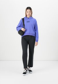 Femme portant un sweat à capuche violet GAP, un pantalon noir et des baskets noir et blanc, tenant un sac à bandoulière noir contre un fond blanc uni.