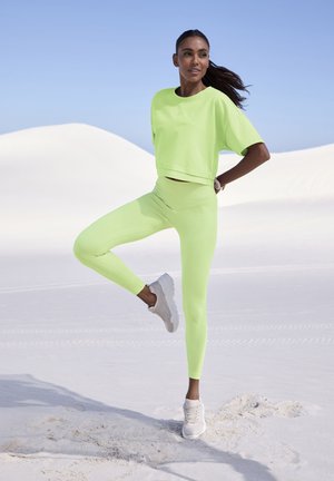 Neongrünes Cropped-Top und passende Leggings aus geripptem Stoff, kombiniert mit hellen Sneakern, vor einem sandigen Wüstenhintergrund.