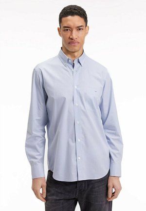 Homme portant une chemise à carreaux bleu clair avec un pantalon noir, se tenant contre un fond blanc uni.
