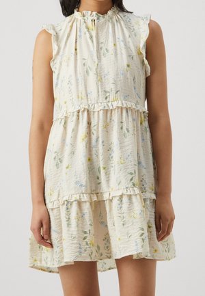 Ærmeløs cremefarvet kjole med lag på lag med flæser og fint mønster af grønne, blå og gule blomster, båret af en stående person.