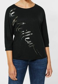 Černé tričko s dlouhým rukávem a kulatým výstřihem, na kterém je diagonálně umístěný metalicky stříbrný text. Lehká tkanina s volným střihem.