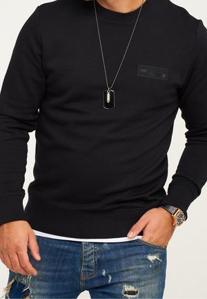 Felpa nera con etichetta testurizzata sul davanti, abbinata a jeans blu strappati. Accessori includono una collana con pendente a targhetta argentata e un orologio.