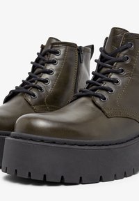 Bottes en cuir vert olive avec une semelle plateforme noire épaisse, un laçage à l'avant et une fermeture éclair sur le côté pour un accès facile. Texture lisse.