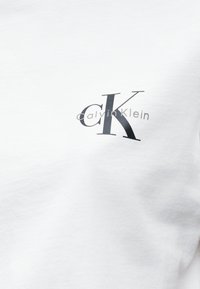 Λευκό ύφασμα με μαύρο λογότυπο αρχικών "cK" και γκρι κείμενο "Calvin Klein" τυπωμένο στην επάνω αριστερή πλευρά ενός ενδύματος.