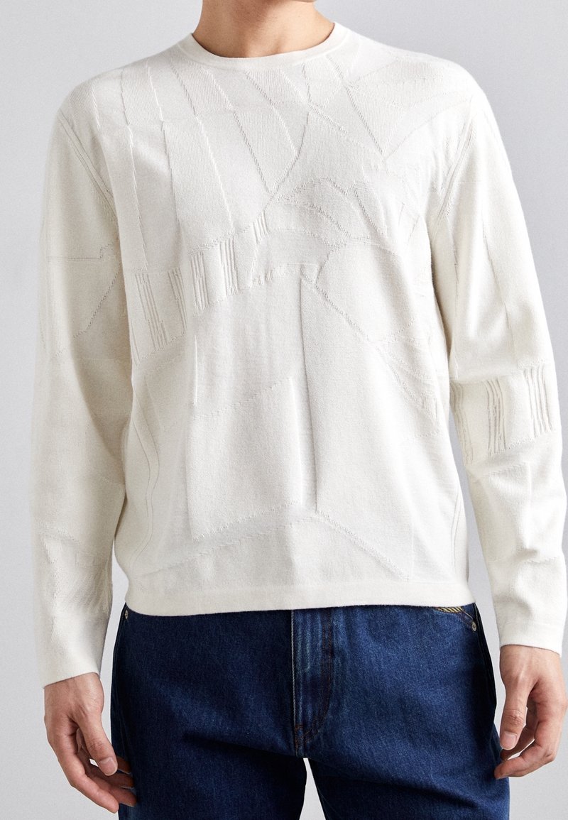 Homme portant un pull blanc texturé à manches longues avec des motifs abstraits et un jean bleu, debout devant un fond neutre.
