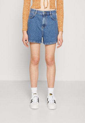 Kvinde model iført mellemblå denimshorts, hvide sokker, hvide sneakers med sorte striber og en kort orange langærmet top.