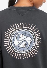 Svart hettegenser med et stort sirkulært grafisk design på baksiden, med et blått og hvitt mønster omgitt av et rosa solstrålemønster.
