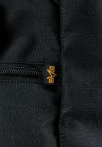 Tela negra texturizada con una cremallera visible y una pequeña etiqueta de logo en naranja. El material parece duradero y bien entrelazado.
