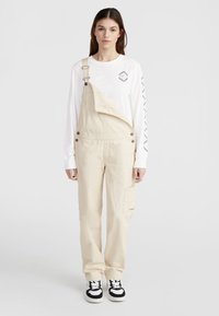 Beige cotton overalls with adjustable straps, front pocket, and button details. Paired with a white long-sleeve shirt and black and white sneakers.