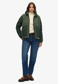 Groene geïsoleerde jas met een textuur van fleece aan de voorkant, twee frontzakken en een ritsluiting. Draag hem over een crèmekleurige coltrui en blauwe jeans.
