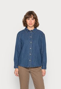 Camisa de ganga com colarinho, mangas longas, fecho frontal por botões e uma cor azul escura com detalhes de costura em contraste.