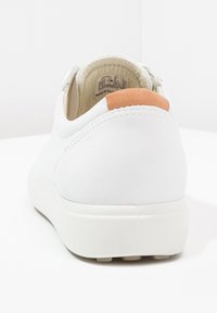 Biały sneaker z grubą podeszwą, widocznymi przeszyciami i jasnobrązową skórzaną pętelką na pięcie, pokazany z tyłu na jednolitym białym tle.