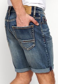 Denim shorts in faded blue, featuring rolled cuffs, standard pockets, and contrasting stitching details. The material has a worn texture.