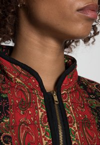 Gequilted jas met een rood paisley patroon, zwarte afwerking en een gouden rits. Heeft een hoge kraag en een zichtbare textuur door het quilten.