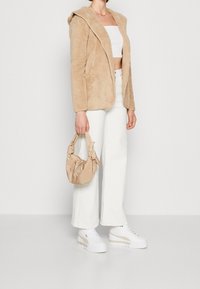 Beżowa kurtka z kapturem z miękkiego materiału, biały crop top, białe jeansy z wysokim stanem oraz brązowa torba z węzłami zamiast uchwytów. Białe sneakersy na platformie.