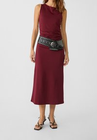 Bordeaux zonder mouwen jurk met rimpeling bij de bodice, gecombineerd met een zwarte gesp met studs. Het schoeisel bestaat uit zwarte sandaaltjes met bandjes.