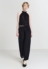 Frau in schwarzem ärmellosem Halterneck-Top und passenden weitgeschnittenen Hosen mit subtilen strukturierten Blumenmustern vor hellem Hintergrund.