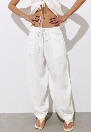 Personne portant un pantalon blanc ample avec un cordon à la taille, un crop top blanc et des talons beiges ouverts à l'avant, debout les mains sur les hanches.