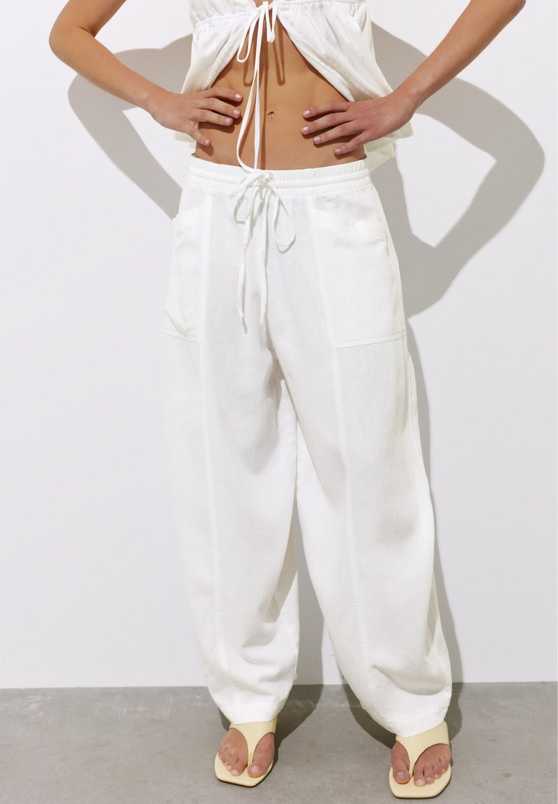 Personne portant un pantalon blanc ample avec un cordon à la taille, un crop top blanc et des talons beiges ouverts à l'avant, debout les mains sur les hanches.