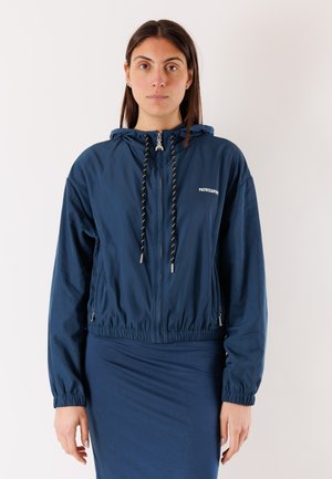 Donna che indossa una giacca con cappuccio blu navy con cerniera, polsini elastici e coulisse, in piedi davanti a uno sfondo chiaro e uniforme.