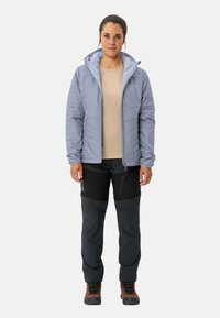 Giacca grigia isolata con cappuccio sopra una maglia beige, abbinata a pantaloni neri con tasche con zip e stivali da trekking marroni.