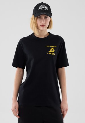 Persona con gorra negra de Los Angeles Lakers y camiseta negra con logo amarillo de Lakers y texto en el pecho, de pie frente a un fondo gris.