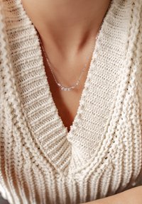 Collier en argent avec des accents en forme de feuille ornés de petites pierres transparentes, porté contre un pull en tricot texturé de couleur crème.