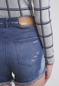 Shorts de mezclilla con dobladillo deshilachado, detalles desgastados y una etiqueta de cuero marrón. Combinados con una blusa de manga larga a cuadros en gris.