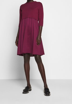 Person trägt ein knielanges, langärmeliges dunkelrosa Kleid und schwarze Loafers mit Quasten und genieteten Sohlen, steht vor einem einfarbigen Hintergrund.