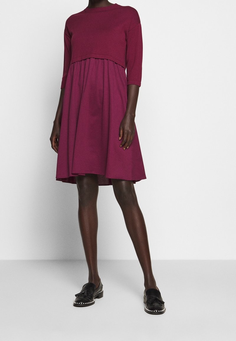 Personne portant une robe rose foncé mi-longue à manches longues et des mocassins noirs avec des pompons et des semelles cloutées, debout devant un fond uni.
