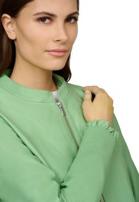 Grüne Lederbomberjacke mit einem runden Kragen, gerippten Bündchen und sichtbarem silbernem Reißverschluss. Glatte Textur mit einer taillierten Silhouette.