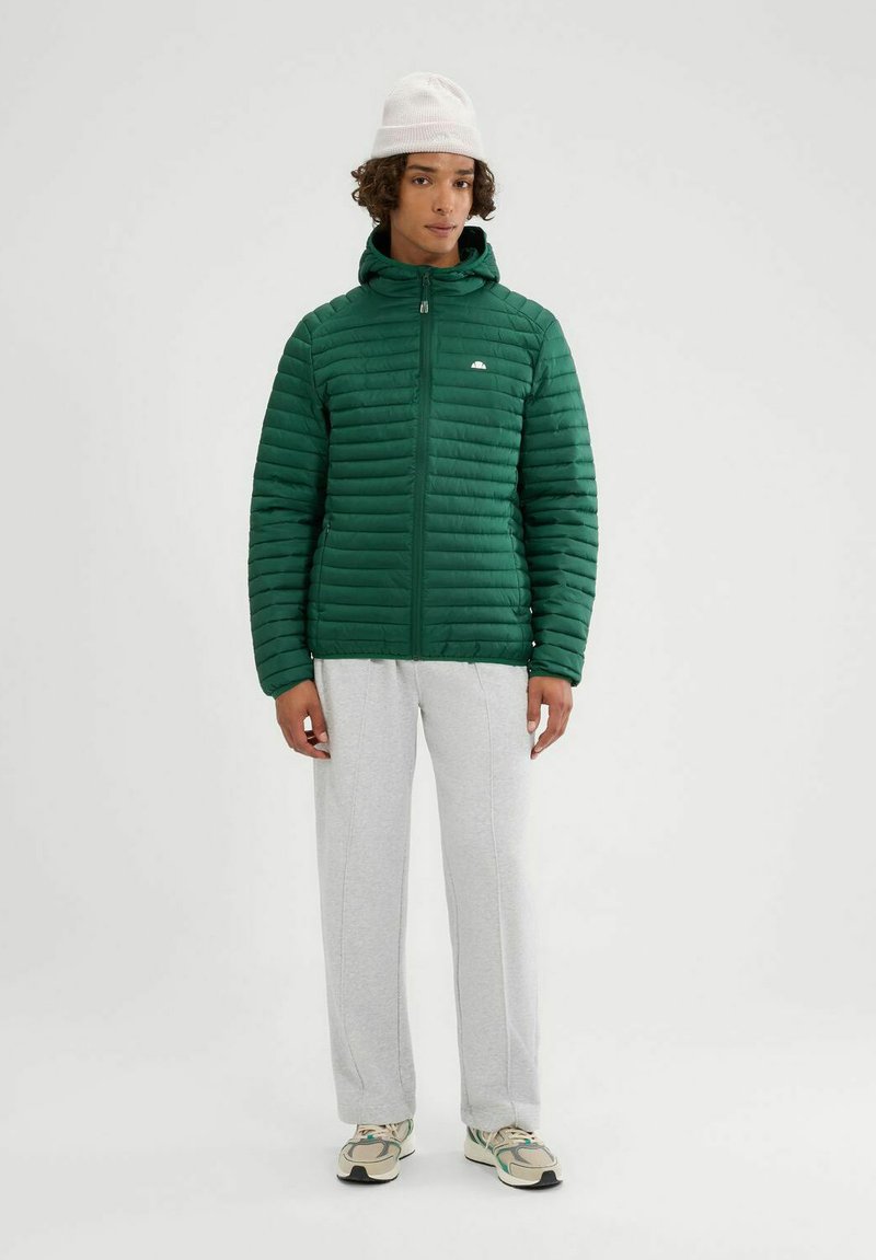 Giacca verde imbottita con chiusura a zip, colletto alto e cuciture orizzontali, abbinata a pantaloni della tuta grigi e un cappello bianco.