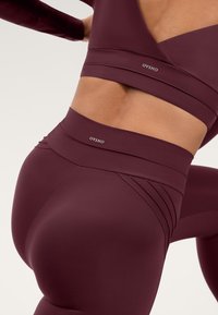 Set athletic de culoare vișinie, format din colanți cu talie înaltă și un tricou crop ajustat. Materialul pare neted, cu detalii subtile ale cusăturilor și accente cu logo.