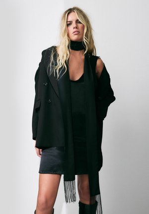 Schwarzer Blazer über einem ärmellosen Kleid aus glattem Stoff. Der Look wird durch einen schwarzen Schal mit Fransen und kniehohen schwarzen Stiefeln ergänzt.