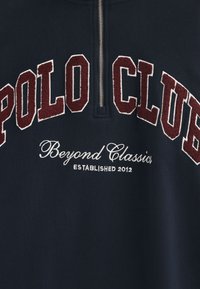 Marinblå sweatshirt med en kvart dragkedja, med upphöjd burgundy-text "POLO CLUB" och vit broderad detalj "Beyond Classics Established 2012."