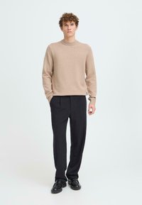 Beige Strickpullover mit Rundhalsausschnitt, langen Ärmeln und gerippten Bündchen, kombiniert mit schwarzen plissierten Hosen und schwarzen Schuhen. Schlichtes Design, weiche Textur.