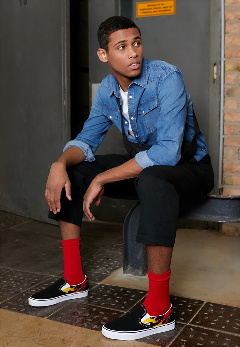 Zapatos negros tipo slip-on con diseño de llamas, suela blanca, combinados con calcetines rojos hasta la altura del tobillo. El modelo lleva una camisa de mezclilla sobre una camiseta blanca y pantalones negros.