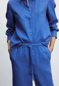 Blauw linnen overhemd met kraag, knopen aan de voorkant en opgerolde mouwen; gecombineerd met bijpassende blauwe broek met trekkoord en zijzakken.