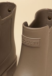 Beige rubber ankle boots with the brand name "Joules" embossed on a patch on the front upper part of the right boot.