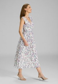 Floralmaxikleid in Weiß mit lila und blauen Mustern. Mit Rüschenträgern und einer taillierten Taille. Kombiniert mit silbernen Riemchenschuhen.