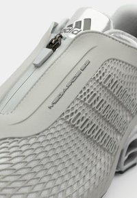 Zapatilla deportiva de color gris claro con una parte superior de malla texturizada, cierre de cremallera y el logo grabado "MEGARIDE 62". Presenta acentos plateados y un diseño elegante.