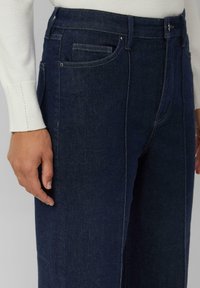 Ciemnoniebieskie dżinsy z prostymi nogawkami, pięcioma kieszeniami i subtelnymi detalami przeszyć. Tkanina wydaje się wytrzymała i teksturowana.