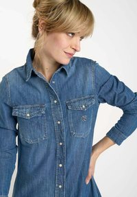 Chemise en denim avec un devant à boutons, deux poches poitrine et un logo brodé. Manches longues avec poignets boutonnés et design classique.