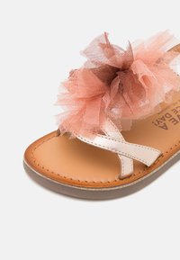 Sandalia con tiras y un upper de metalizado rosa dorado, plantilla en tono marrón claro y un gran detalle de flor de tul rosa en la parte delantera. Diseño de suela plana.