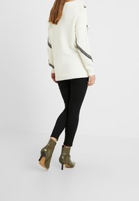Suéter de punto blanco con detalle a cuadros en negro, combinado con leggings negros y botas de tobillo verde oliva con punta afilada y tacón de aguja.