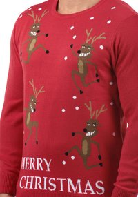 Pull de Noël rouge avec des motifs ludiques de rennes et des flocons de neige blancs. Le texte "JOYEUX NOËL" est inscrit en blanc en bas.