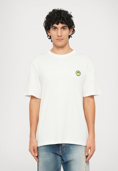 Camiseta de algodón blanca con mangas cortas y cuello redondo. Presenta un pequeño parche de cara sonriente amarilla en el área del pecho izquierdo.
