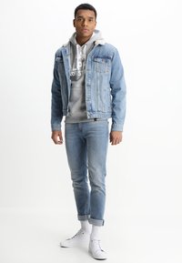Chaqueta de denim azul clara con un cuello de forro polar blanco, sudadera gris con logo, vaqueros azul claros y zapatillas blancas con puños enrollados y calcetines blancos.