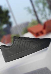 Baskets basses noires avec bandes cousues et lacets, montrées en extérieur sur une surface blanche avec un arrière-plan flou.